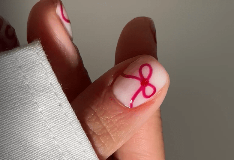 A hand with white-painted nails, of which the nail of the thumb is decorated with a pink bow.