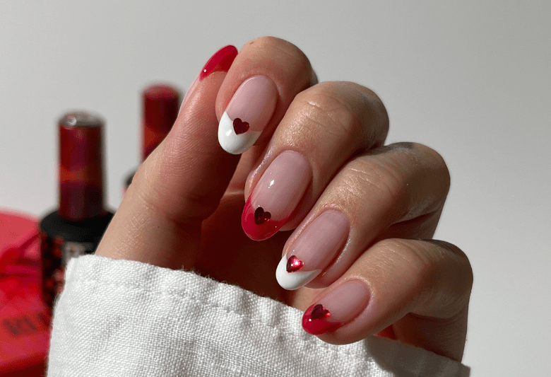 A hand with pink nails featuring a red and white French manicure with heart-shaped decorations.