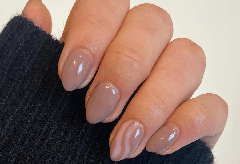 Hand with almond-shaped nails painted in a shiny beige color, with the ring fingernail showing a subtle marble pattern, against a dark sweater.