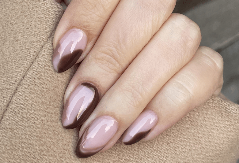 A hand with shiny nails in a light pink color with dark brown accents, worn against a beige fabric.