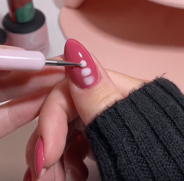 Close-up of a hand decorating a nail with pink polish with a dot pattern in light pink with a nail art tool.