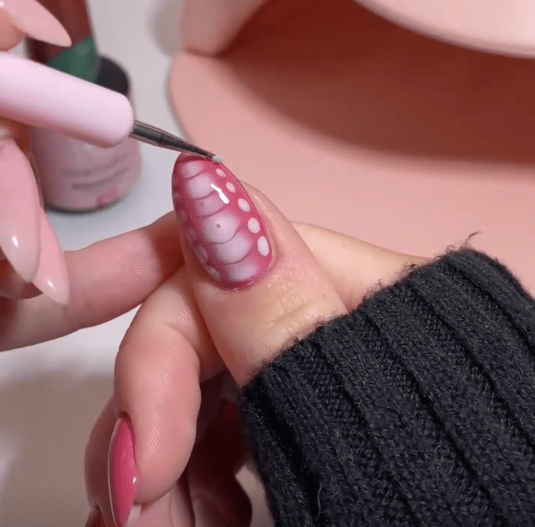 A person is engaged in detailed decoration of a nail with a pink and white pattern, using a brush.