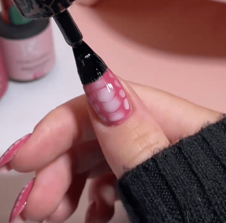 A hand with pink nails decorated with a bubble design, while a black nail polish brush is applied over the thumb.