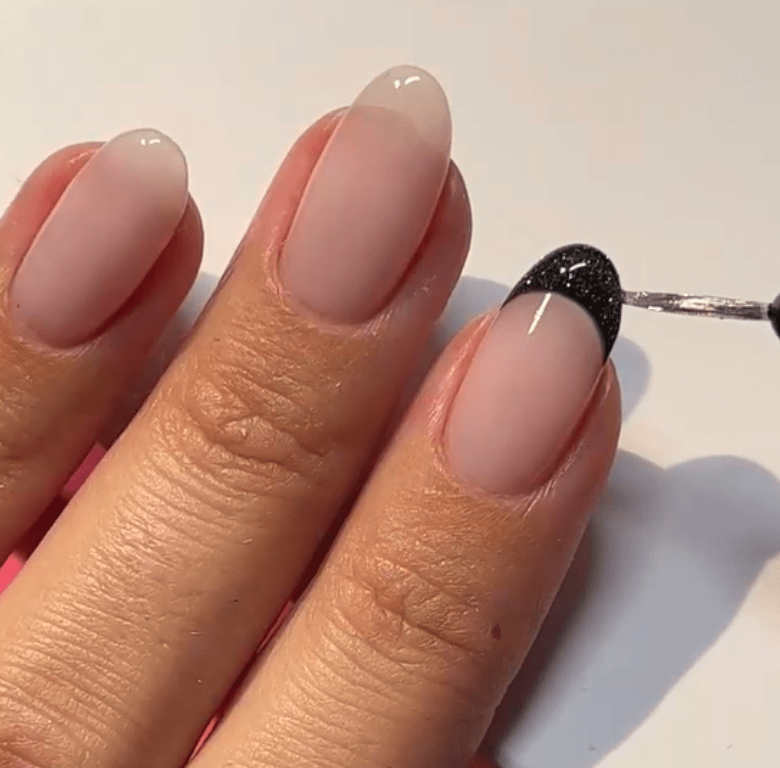 A hand with three fingers, where a black glittering nail polish pattern is applied to the tip on the middle finger.