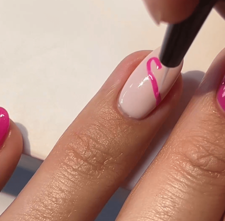 A hand holding a nail with a light pink background varnish and a pink design pattern applied with a brush.
