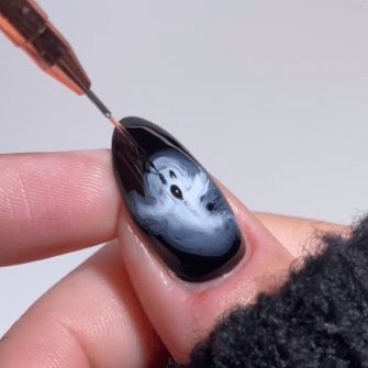 A close-up of a hand with a black painted nail, on which a small white ghost is painted with a fine brush.