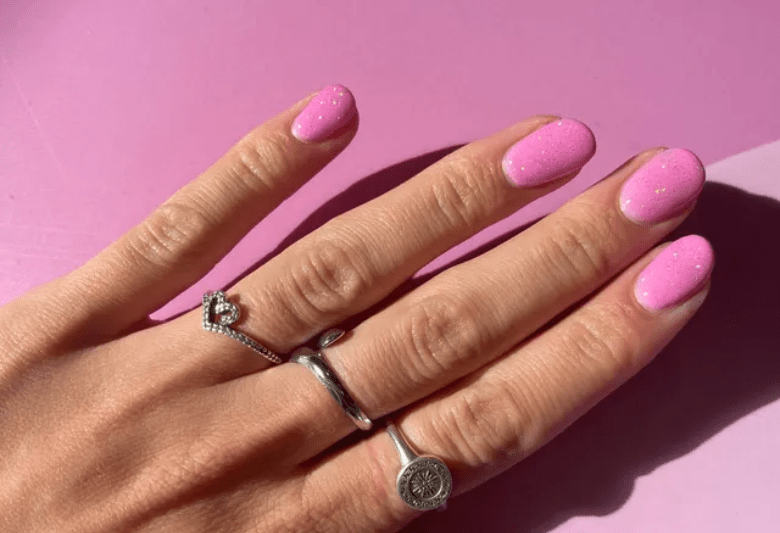 A hand with pink glitter nail polish and three silver rings, against a pink background.