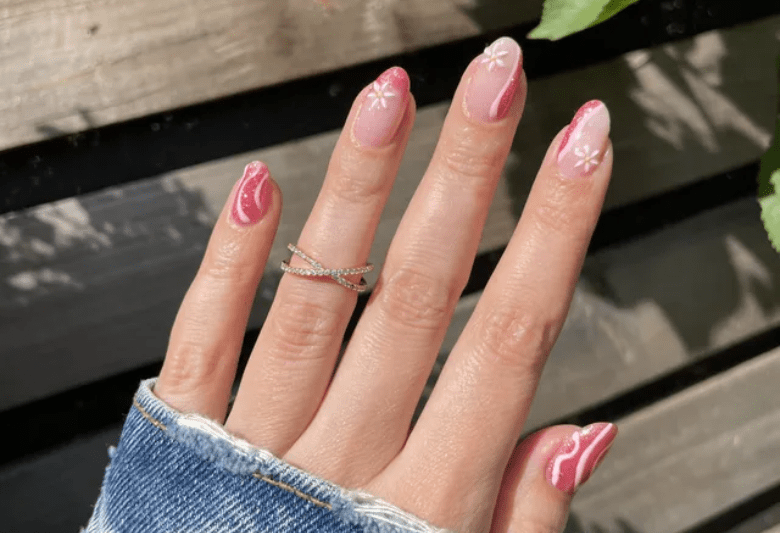 Hand holding pink nail polish and white flowers, worn in a ripped jeans sleeve with a silver ring against a wooden background.
