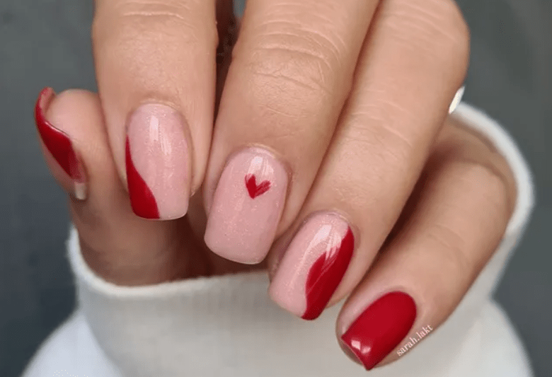 Close-up of a hand with pink nail polish and red accents, including a small red heart on the middle fingernail.