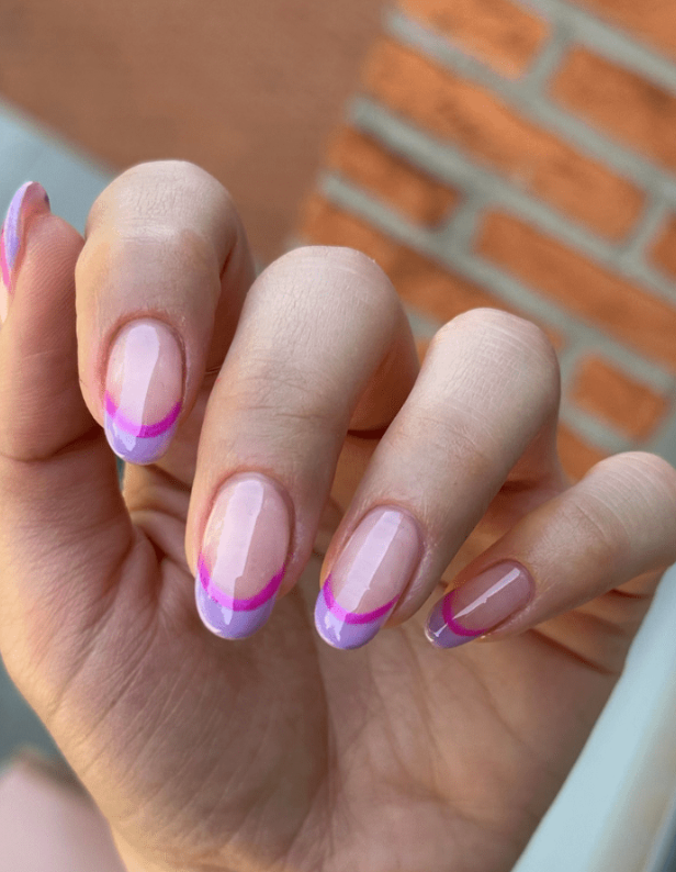 Close-up of a hand with a nail design in pastel colors, consisting of purple and pink edges against a neutral base, with a brick wall in the background.