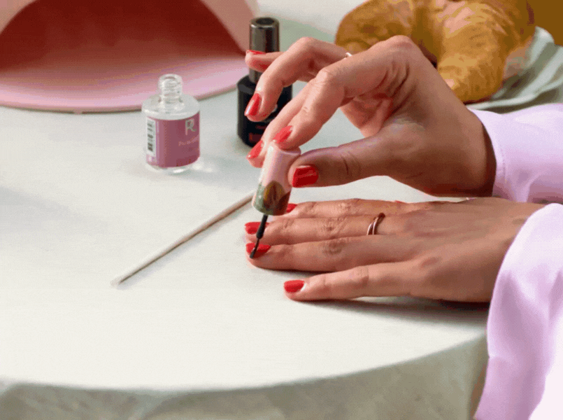 Person painting nails red at a table with nail polish bottles and a croissant.