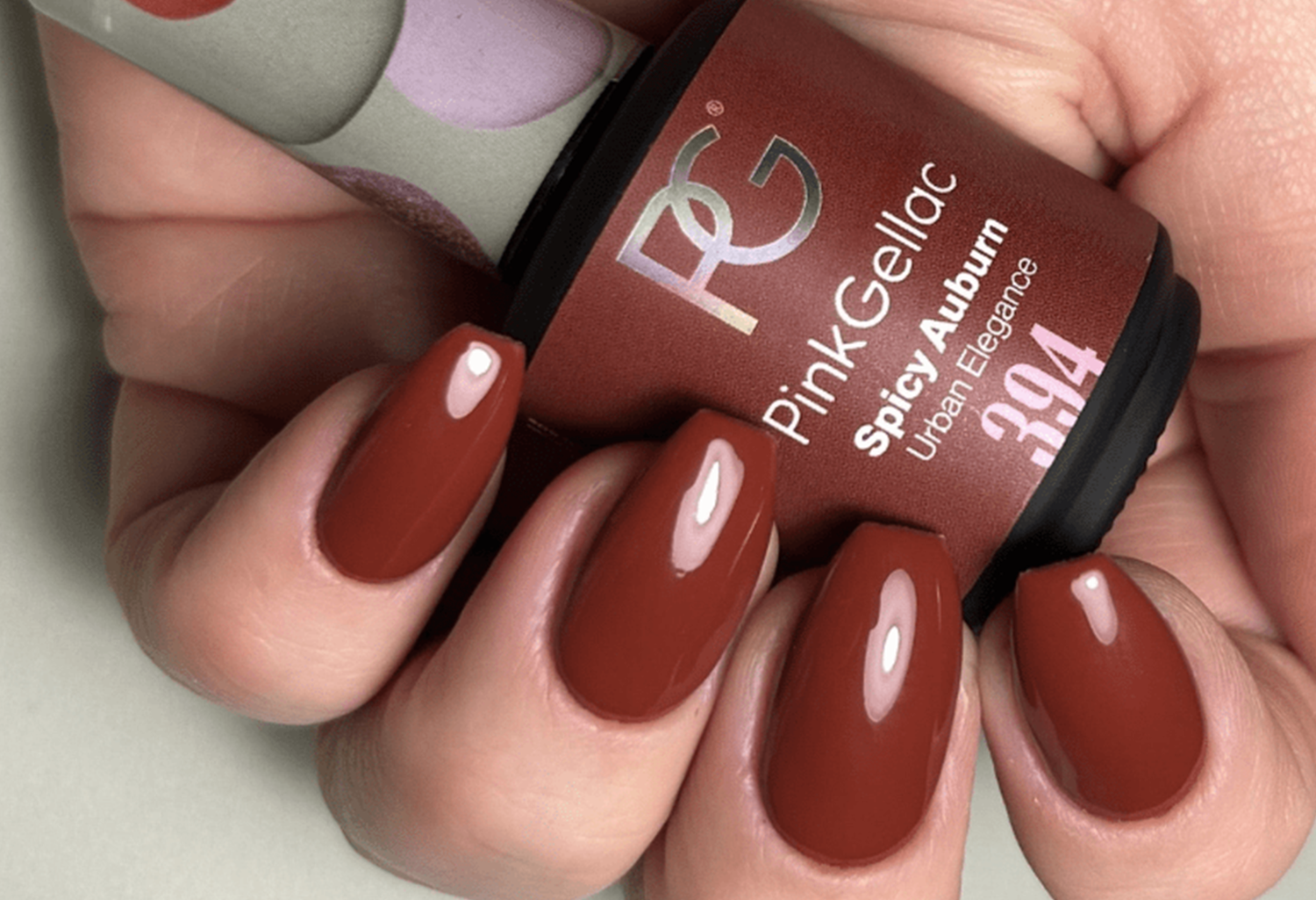 A hand with brown-red nail polish holds a bottle of Pink Gellac Spicy Auburn.