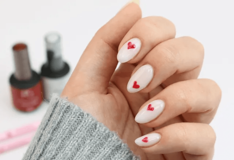 Close-up of a hand with pale pink nails decorated with small red hearts, nail polish bottles can be seen in the background.