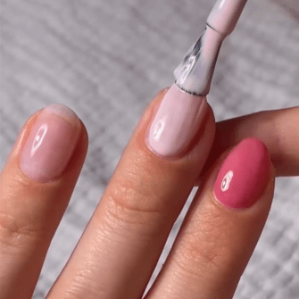 A hand with perfectly manicured nails, two of which are painted pink; A nail is painted light pink by a brush.
