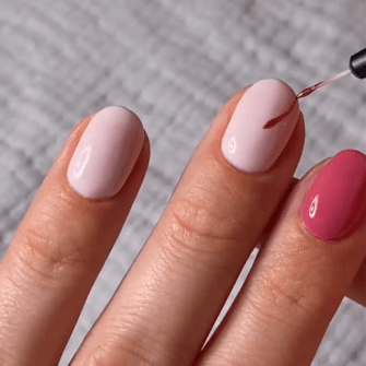 A hand with stylized nails in pale pink and fuchsia, on which varnish is applied.