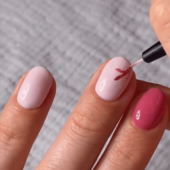 A hand with pink painted nails, applying a design with a darker pink nail polish brush.