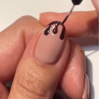 A close-up of a hand applying a dark nail polish in a dripping pattern to a neutrally painted nail.