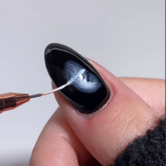 A black nail with detailed paintwork in white and gray tones, where a brush applies a fine pattern.
