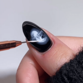 Close-up of a hand with a nail artist painting elegant white patterns on a black nail with a fine brush.
