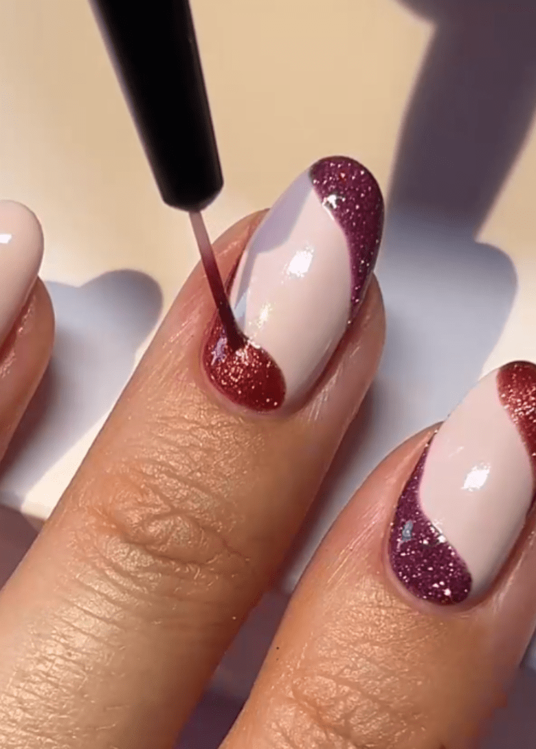 Close-up of a hand with painted nails, using a brush to apply a burgundy glitter polish to a nail with a pink base and purple glitter edges.