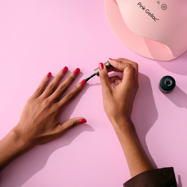 Someone paints pink nail polish on a hand against a pink background, next to a bottle of nail polish and a nail lamp.
