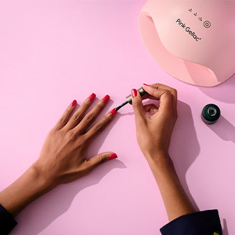 A person paints pink nail polish on her nails, using a jar of nail polish and a UV lamp on a pink surface.