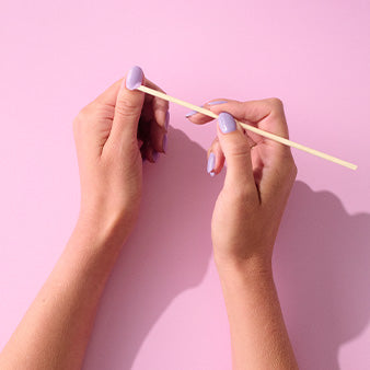 Two hands with purple nail polish hold a wooden stick against a pink background.