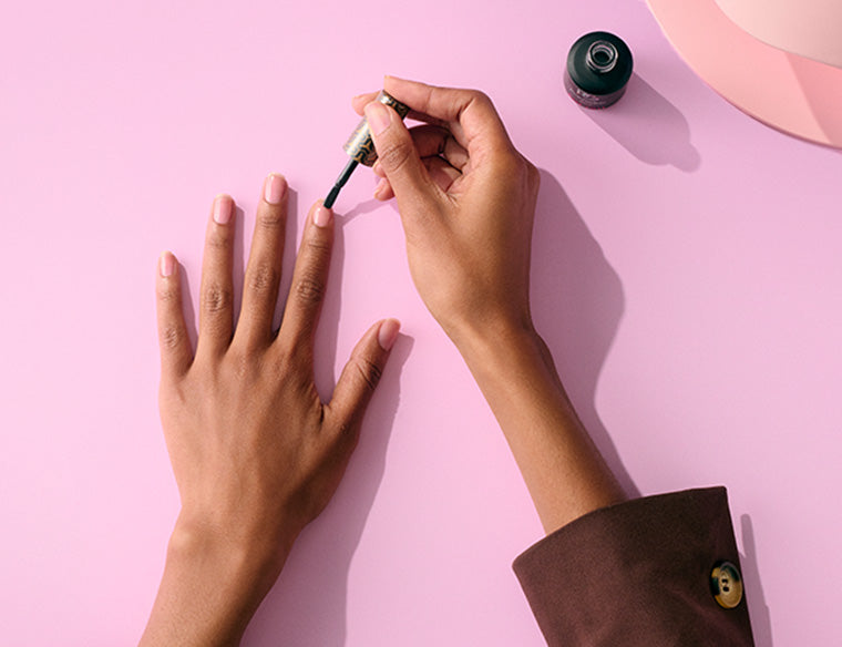 A person paints his nails with paint on a pale pink background.