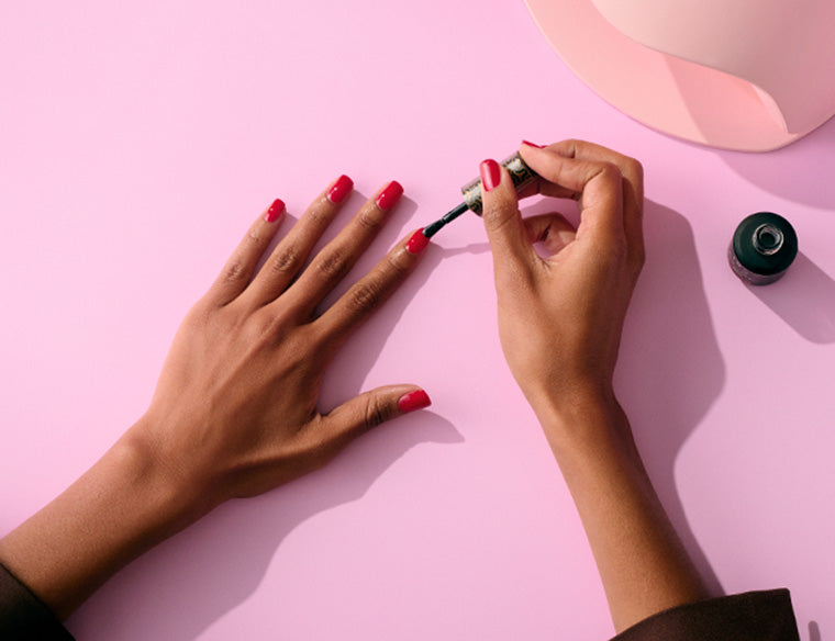 Hands applying dark pink nail polish on a light pink background.
