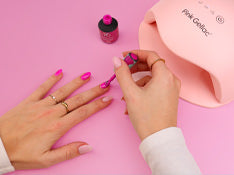 A person paints nails with pink nail polish on a pink background, next to a nail dryer.