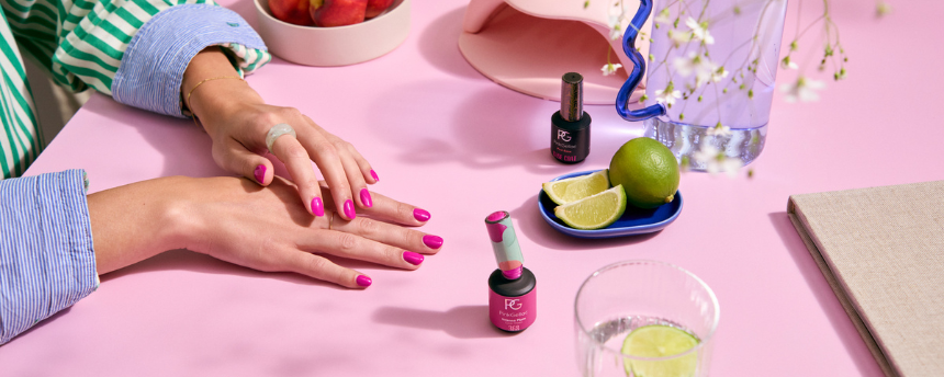 Hands with bright pink nails on pink table, two nail polish bottles, lime wedges, and a glass of water.