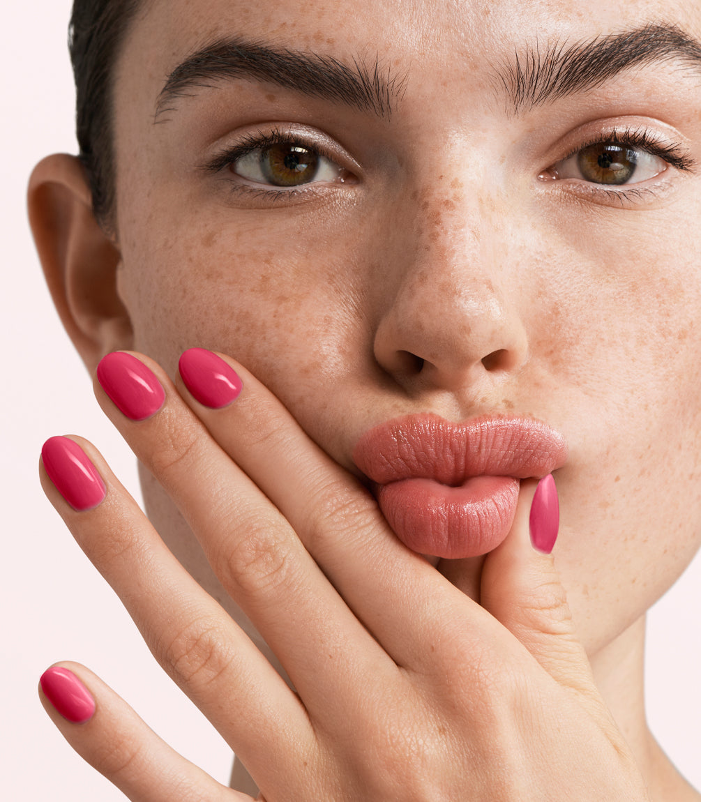 Close-up of a freckled face, with the person pursed her lips and holding her hand with bright pink nail polish under her mouth.