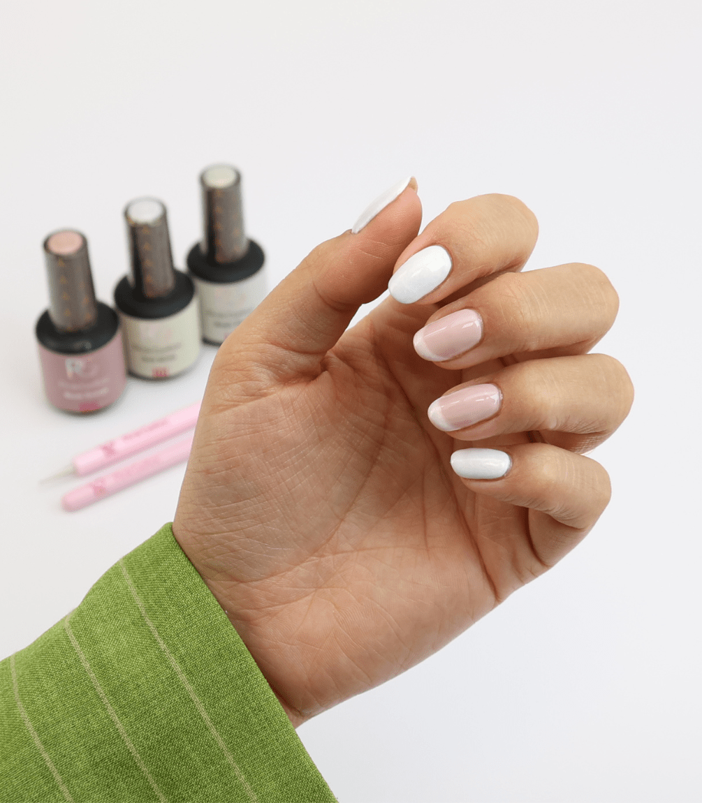 Hand holding pastel pink and white nails, in the background three bottles of nail polish and pink nail art brushes, a green sleeve visible.