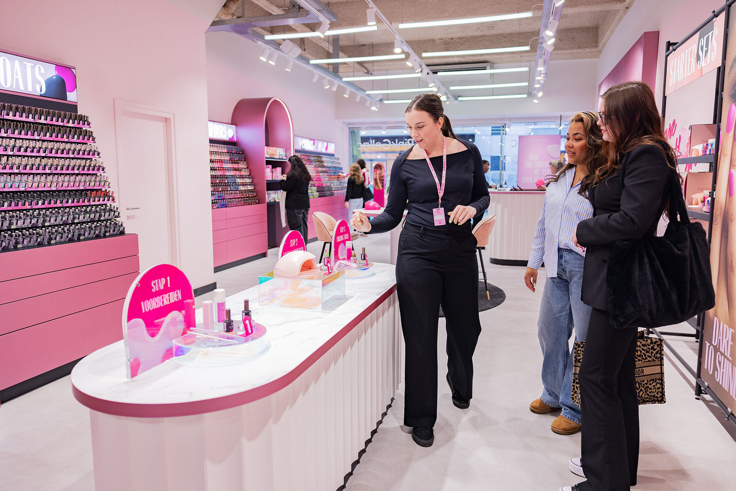 A shop assistant explains something to two customers at a counter in a brightly colored cosmetics store with nail polish displays.