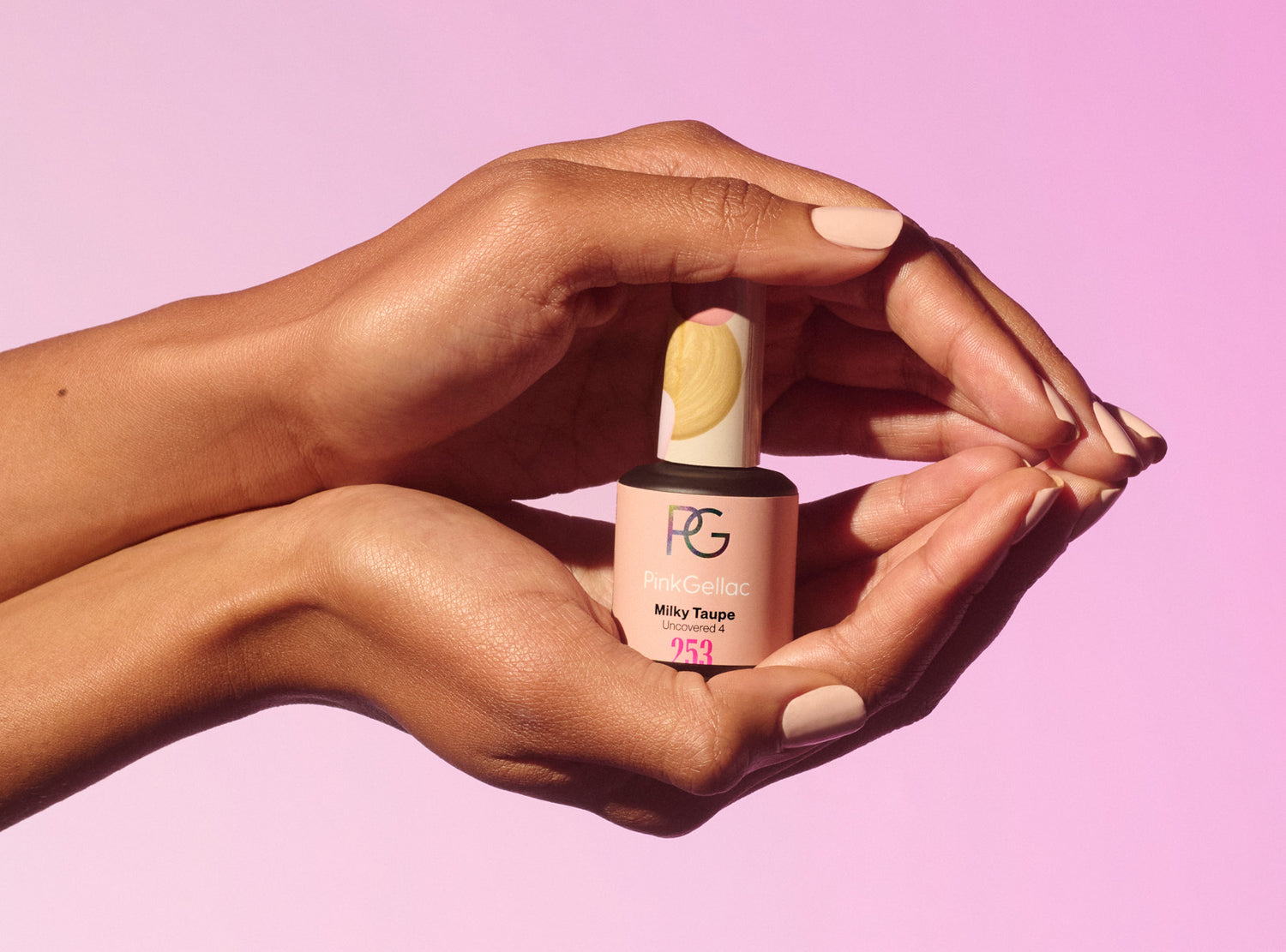 Two hands with light pink painted nails hold a bottle of nail polish from the brand Pink Gellac, color "Milky Taupe", against a pink background.