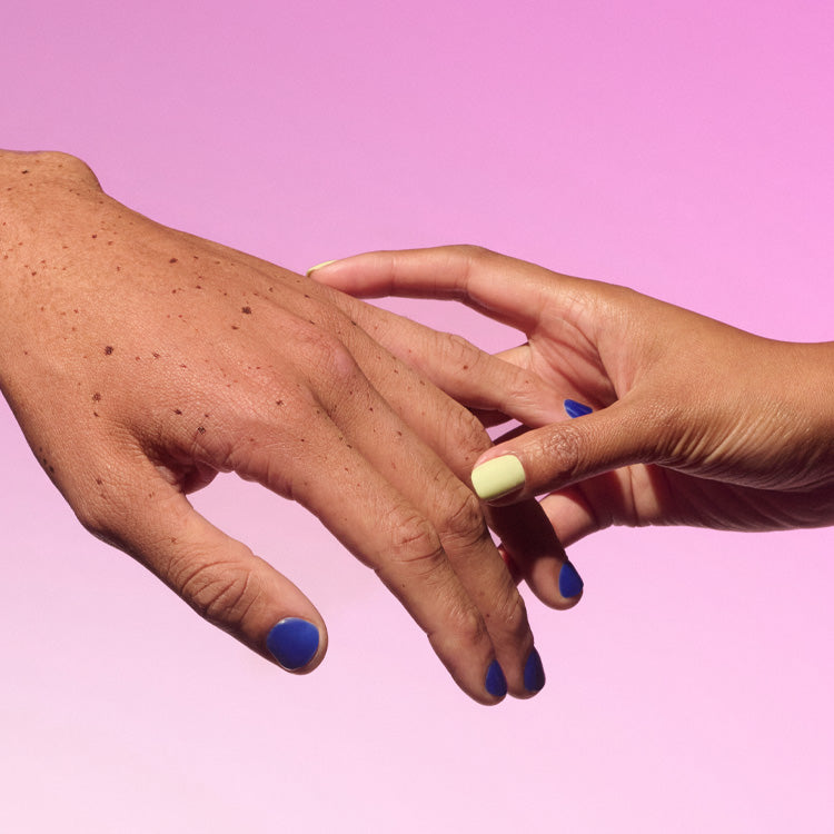 Two hands with colorful nail polish, one hand gently holding the other, and a pink background.