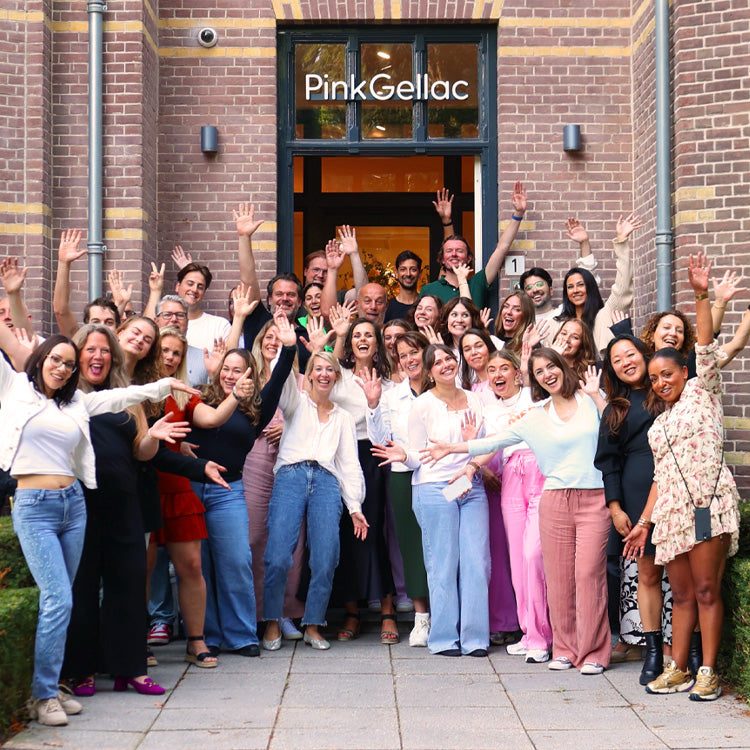 A large group of people happily pose with their arms in the air in front of the entrance of a building with a sign with "Pink Gellac" on it.