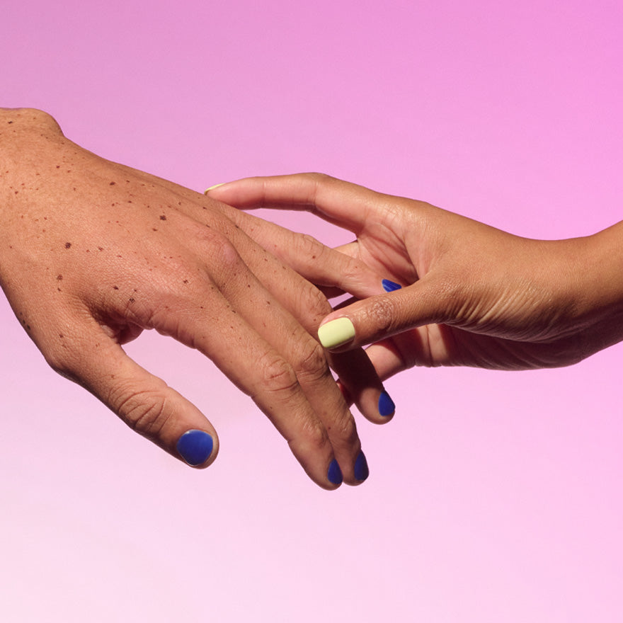 Two hands gently touching each other against a pink background, with blue and yellow painted nails.