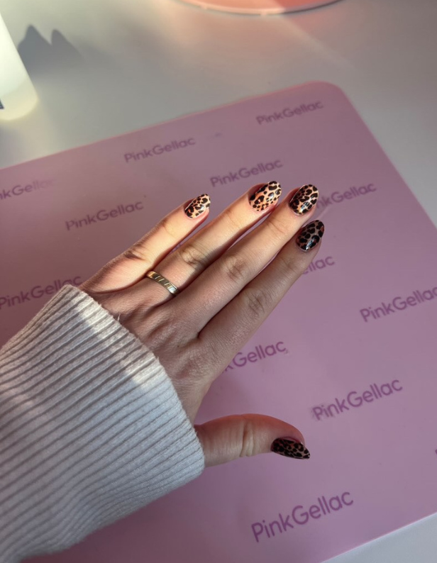 Hand with leopard print nail design and a gold ring, resting on a pink mat with the logo "Pink Gellac".