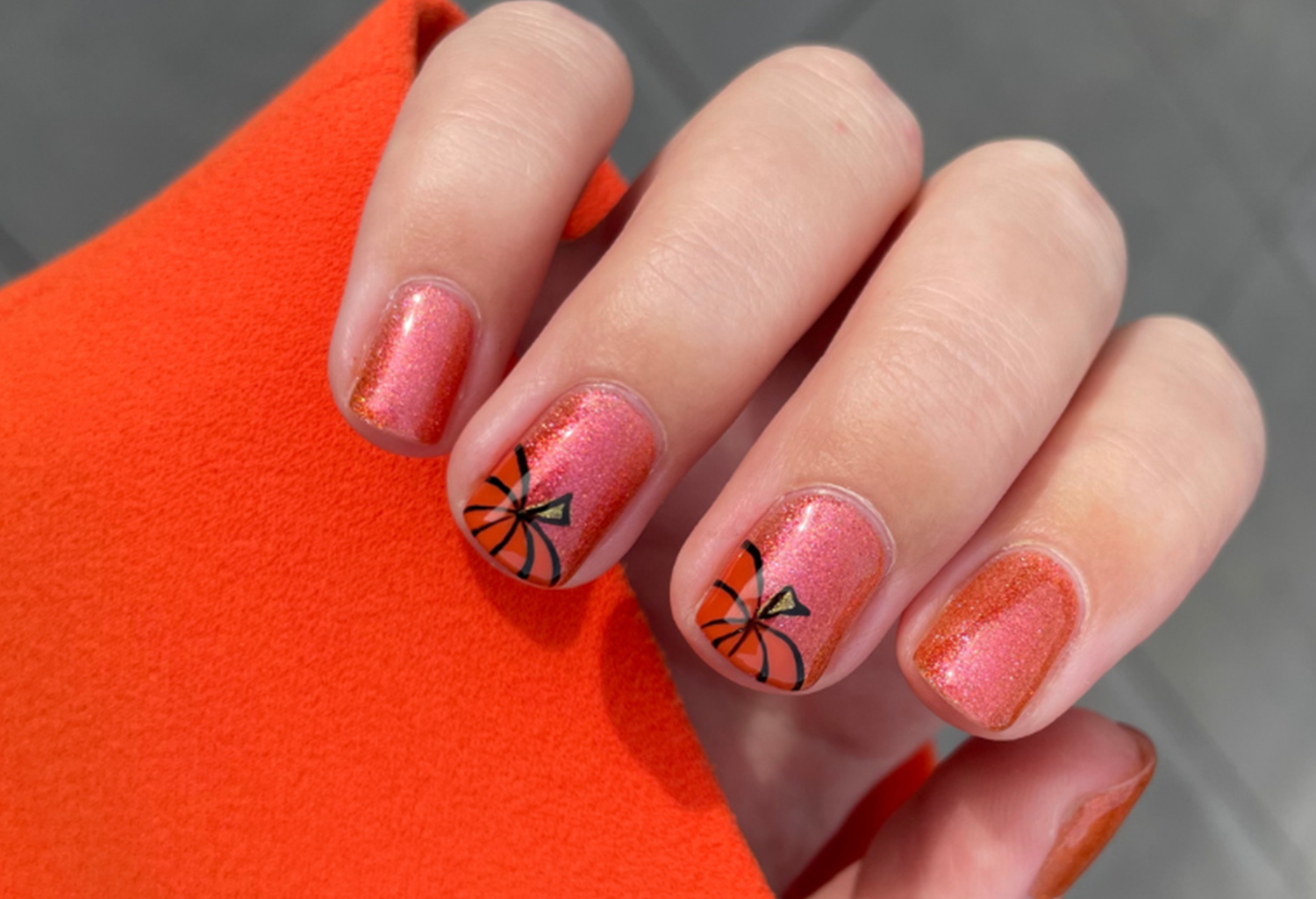 Nails with pink glitter polish and pumpkin designs, hand resting on orange fabric.