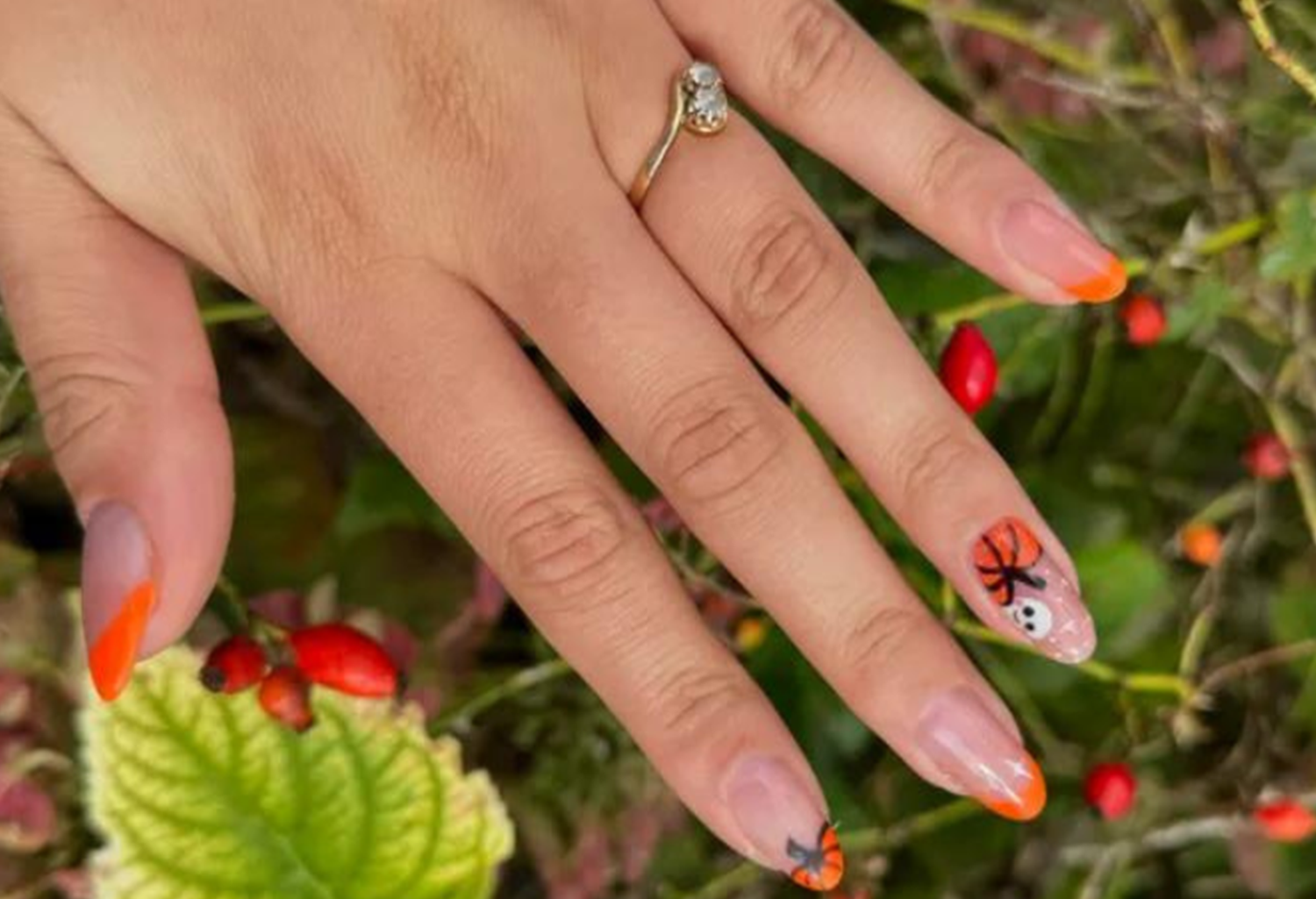 Hand with orange Halloween nail art, including spiders and pumpkins, against a background of green leaves and red berries.
