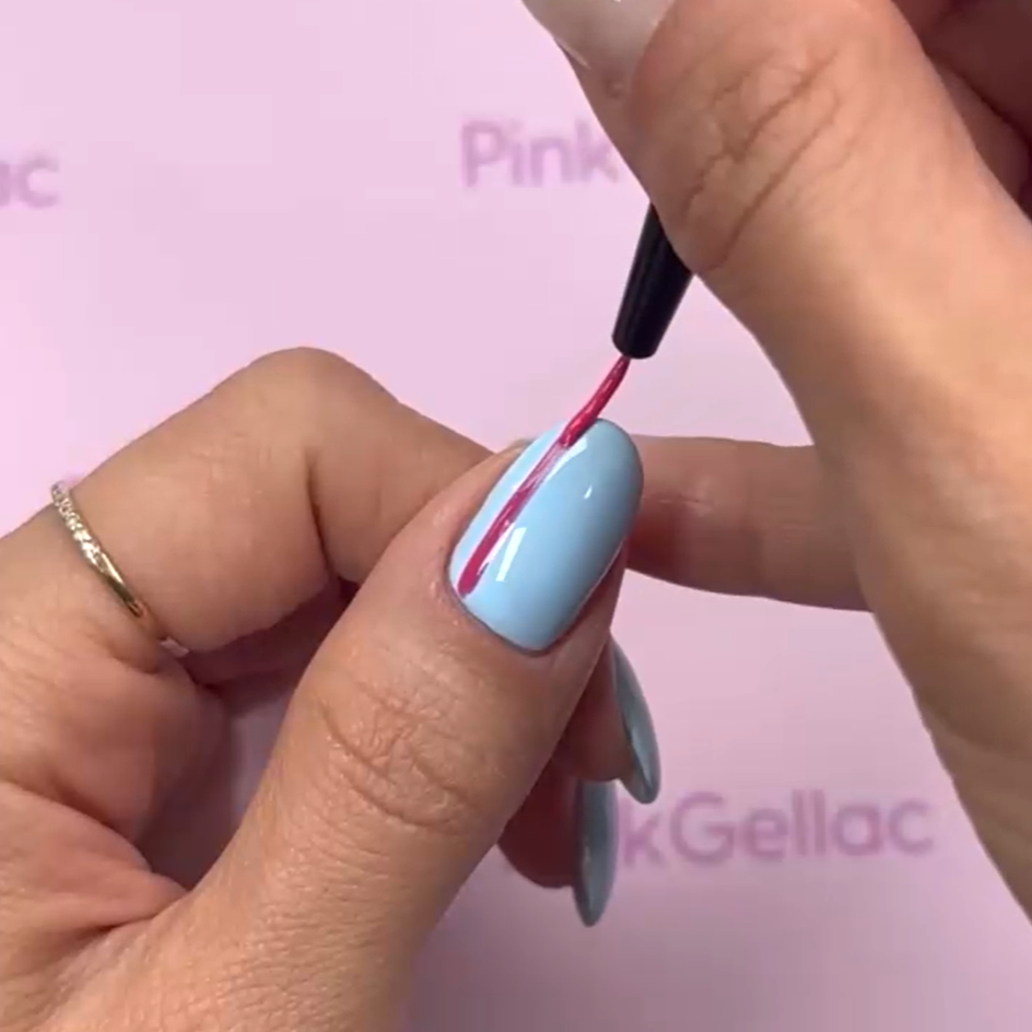 A hand with a blue nail gets a pink stripe painted with a thin brush, against a pink background.