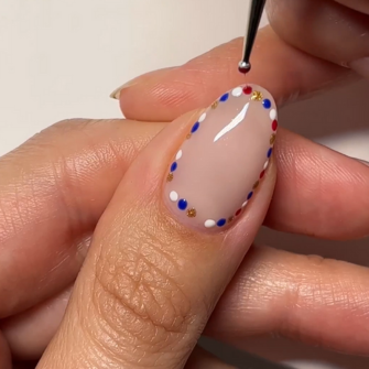 A hand with a nail decorated with red, white, blue and gold dots along the edge. A brush adds details.