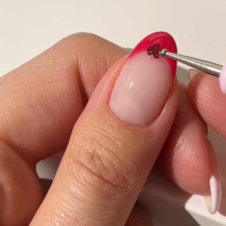 Close-up of a hand painting a red heart on a nail with pink nail polish.