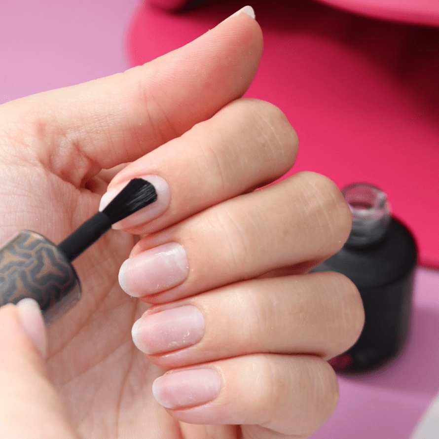 A close-up of a hand applying nail polish to nails, with a pink background.