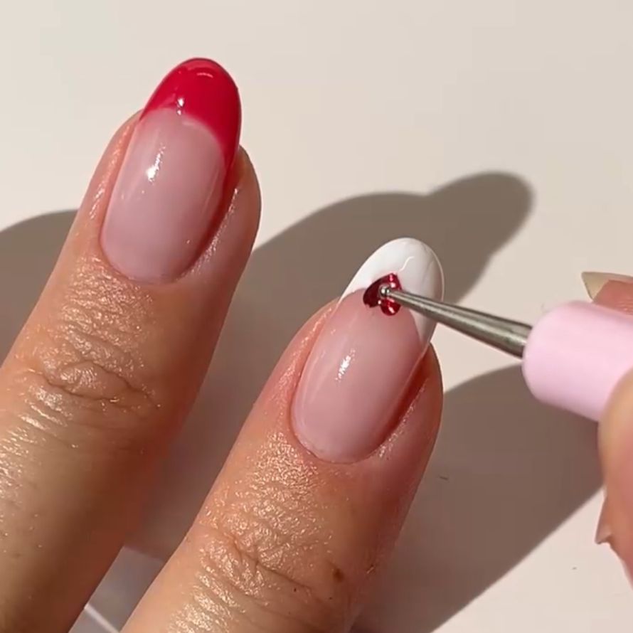 Close-up of two fingers with nail polish; One nail has a red French manicure, while the other is white with a red heart that is applied with a dotting tool.