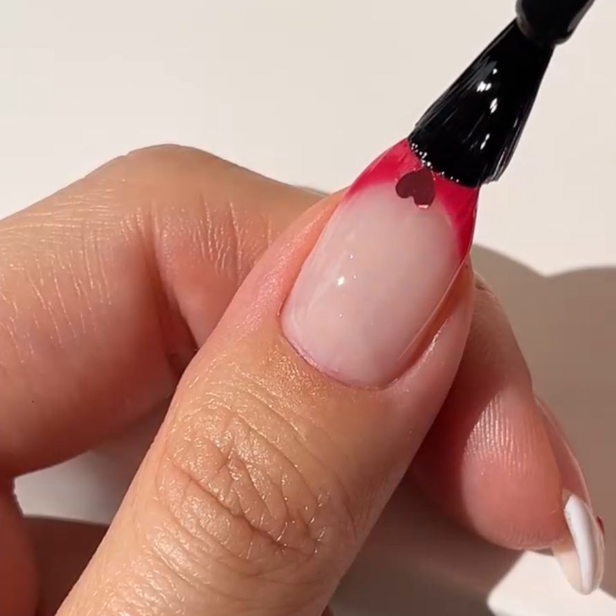 A nail is painted with a brush with pink nail polish and a small heart near the edge.