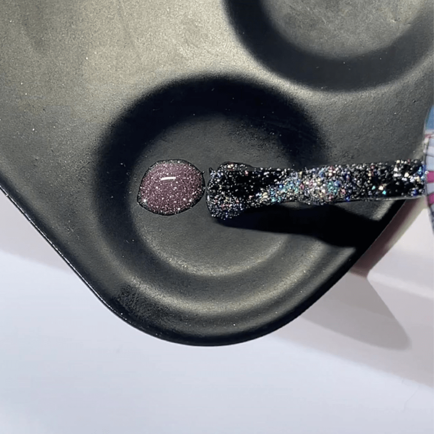 Brush with glittering nail polish above a black pallet with a purple drop.