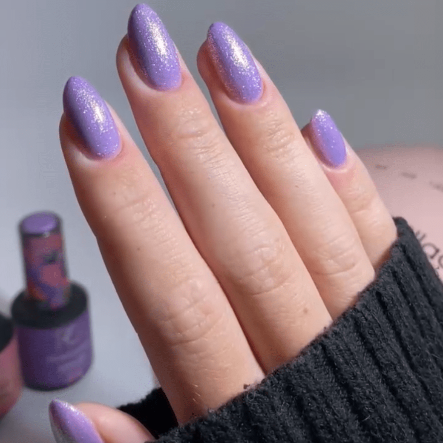 A hand with bright purple, shimmering nail polish on almond-shaped nails, next to bottles of nail polish.