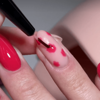 Nail design where a brush applies red dots to a nude-colored nail, next to a bright red painted nail.